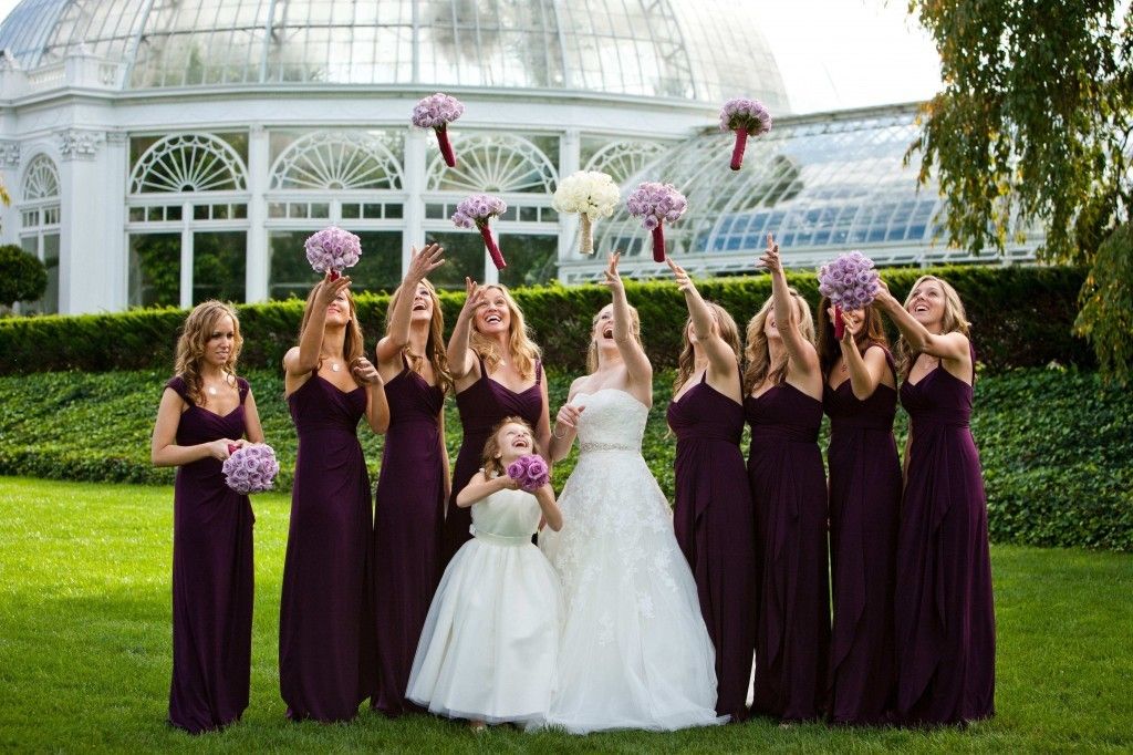 Bridesmaids in deep plum dresses throw bouquets of lavender roses. 